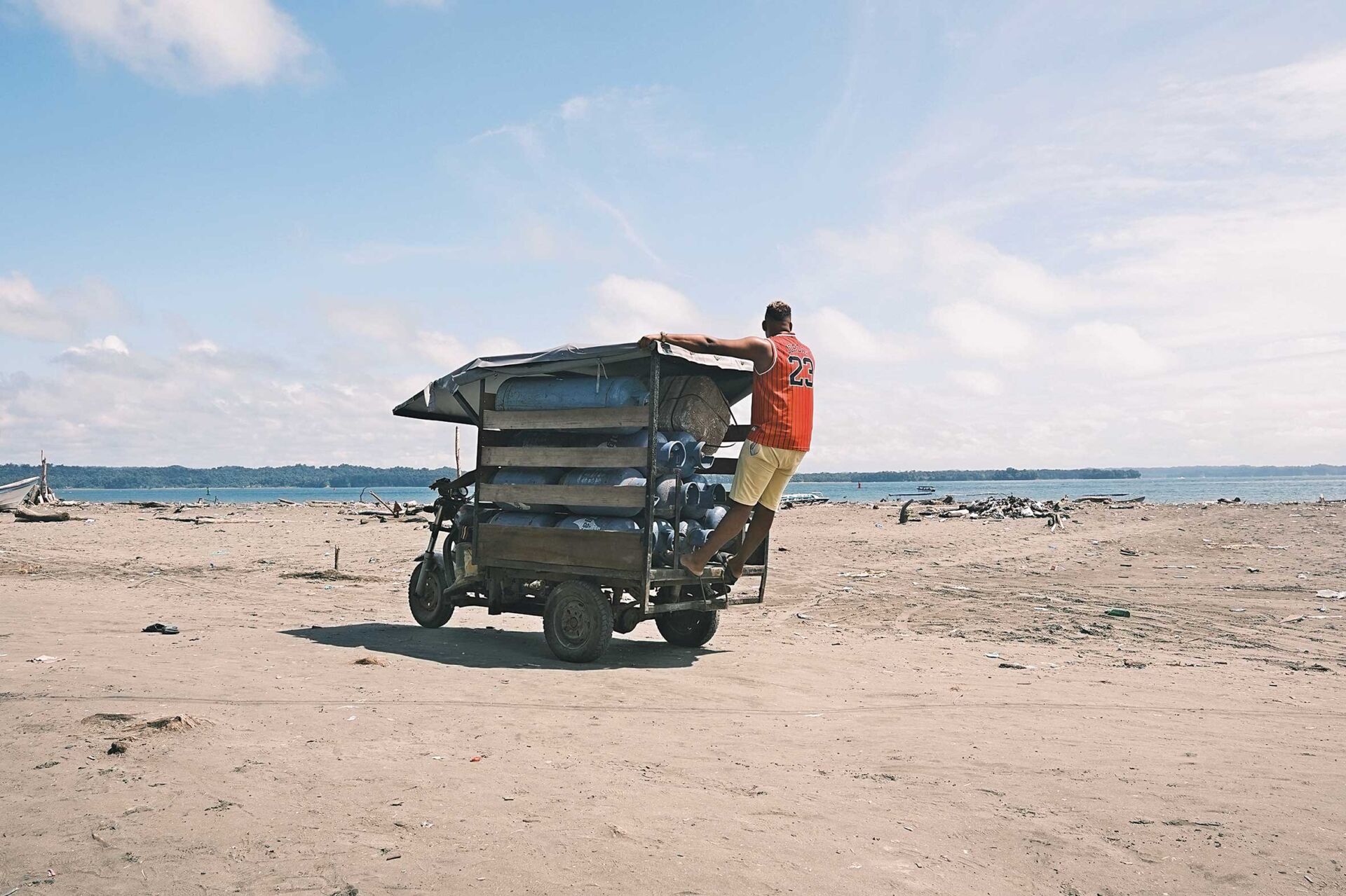 Cotidianidad frente al mar Pacífico. Un hombre se balancea sobre un motocamión cargado de timbos de gas cerca del muelle de Juanchaco, Buenaventura, donde la vida fluye entre lanchas, transeúntes y las labores diarias. Foto de María Andrea Parra.