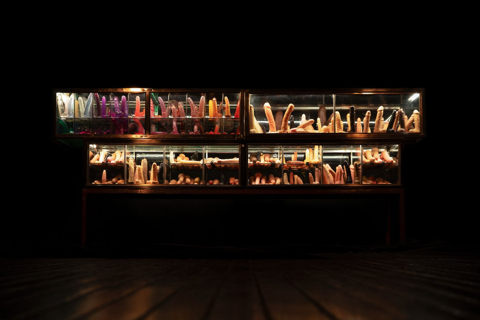 Sin título, 2022-2024, de la serie Ensayo sobre lo oculto y lo revelado de Leonel Castañeda. Instalación de vitrinas metálicas, dildos usados recolectados en mercados callejeros e iluminación LED. La palabra dildo —según una teoría— proviene del italiano diletto, que significa deleite o entretenimiento. Ese deleite aparece trastocado en las rutas del rebusque, en la economía de los residuos. En lugares como las carreras Séptima y Décima, o la calle 18 entre las carreras Séptima y Quinta, en Bogotá, Leonel Castañeda deambula, observa, escoge y recolecta objetos que alguna vez fueron prótesis del deseo, extensiones del cuerpo. En su obra, los acumula y los dispone como cuerpos en sí mismos que fueron usados y gastados, y que ahora han quedado expuestos a la mirada entre la repulsión y la fascinación. El gesto de Castañeda es arqueológico e irónico, y también es estético: nos obliga a mirar lo que normalmente se oculta, lo que provoca vergüenza o asco. Nos enfrenta a una topografía del placer descartado, a una puesta en escena donde el consumo íntimo se transforma en pregunta pública.