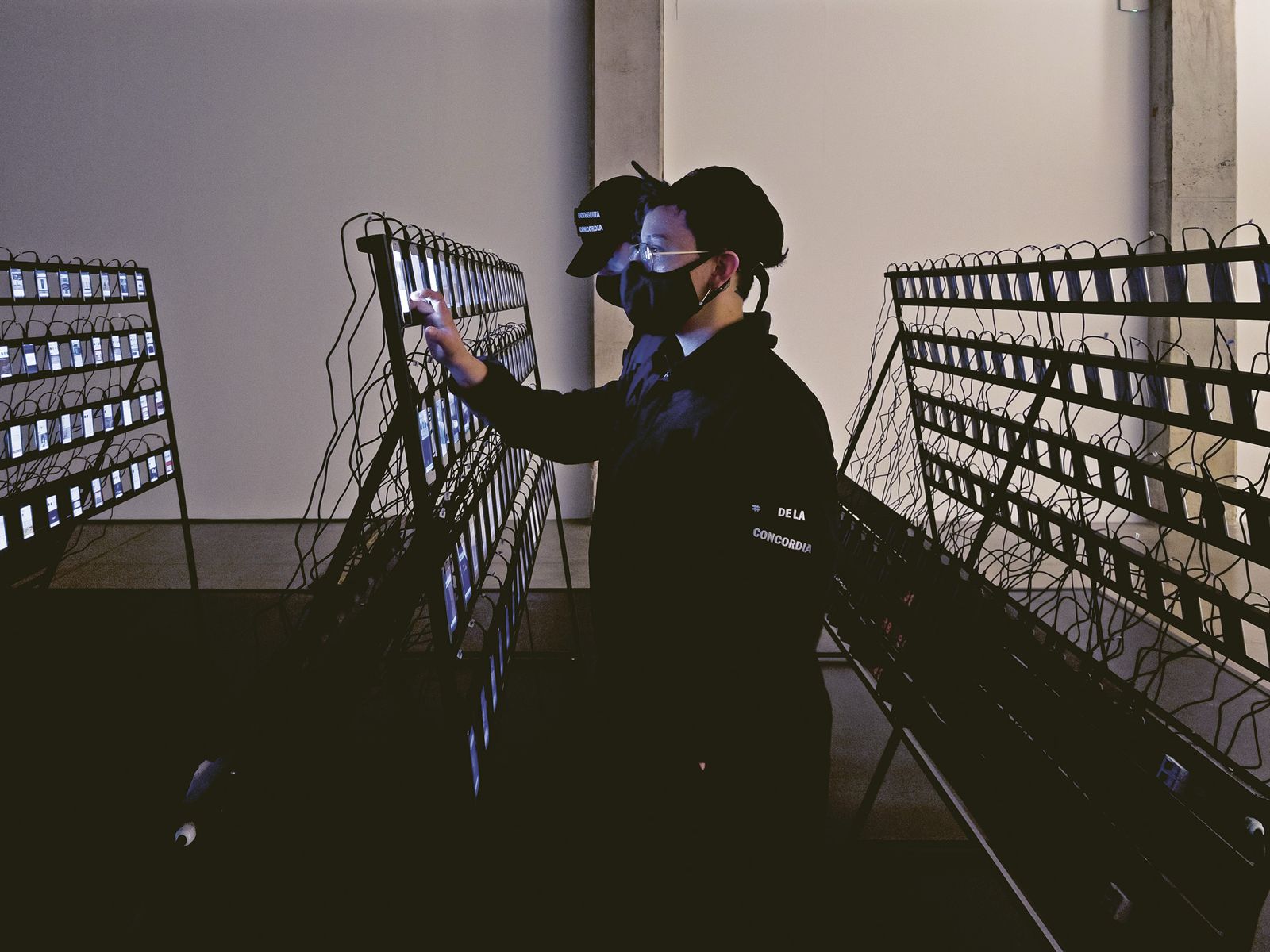 La bodeguita de La Concordia de Juan Obando, 2021. Instalación de trescientos teléfonos móviles, dos computadores de escritorio, dos operarios, mesas y plataforma. «En años recientes, la actividad participativa en internet ha estado ligada a la operación fantasmagórica de redes masivas y automatizadas llamadas granjas de clics. Paralelamente, nuestra relación con la pantalla se ha tornado íntima, personal e incluso espiritual. A través de ella navegamos un cosmos plano, compuesto de imágenes, emojis e íconos, en el que encontramos los materiales necesarios para construir nuestras proyecciones. Aún así, la comprensión colectiva de las operaciones y los sistemas que sostienen esta superficie luminosa es cada vez más opaca. Estimulados por una promesa de viralización, nos desdoblamos como extras y protagonistas de una historia que cabe en nuestros bolsillos pero que poco a poco se está quedando sin batería», dice el artista. Foto de Nicolás Consuegra.