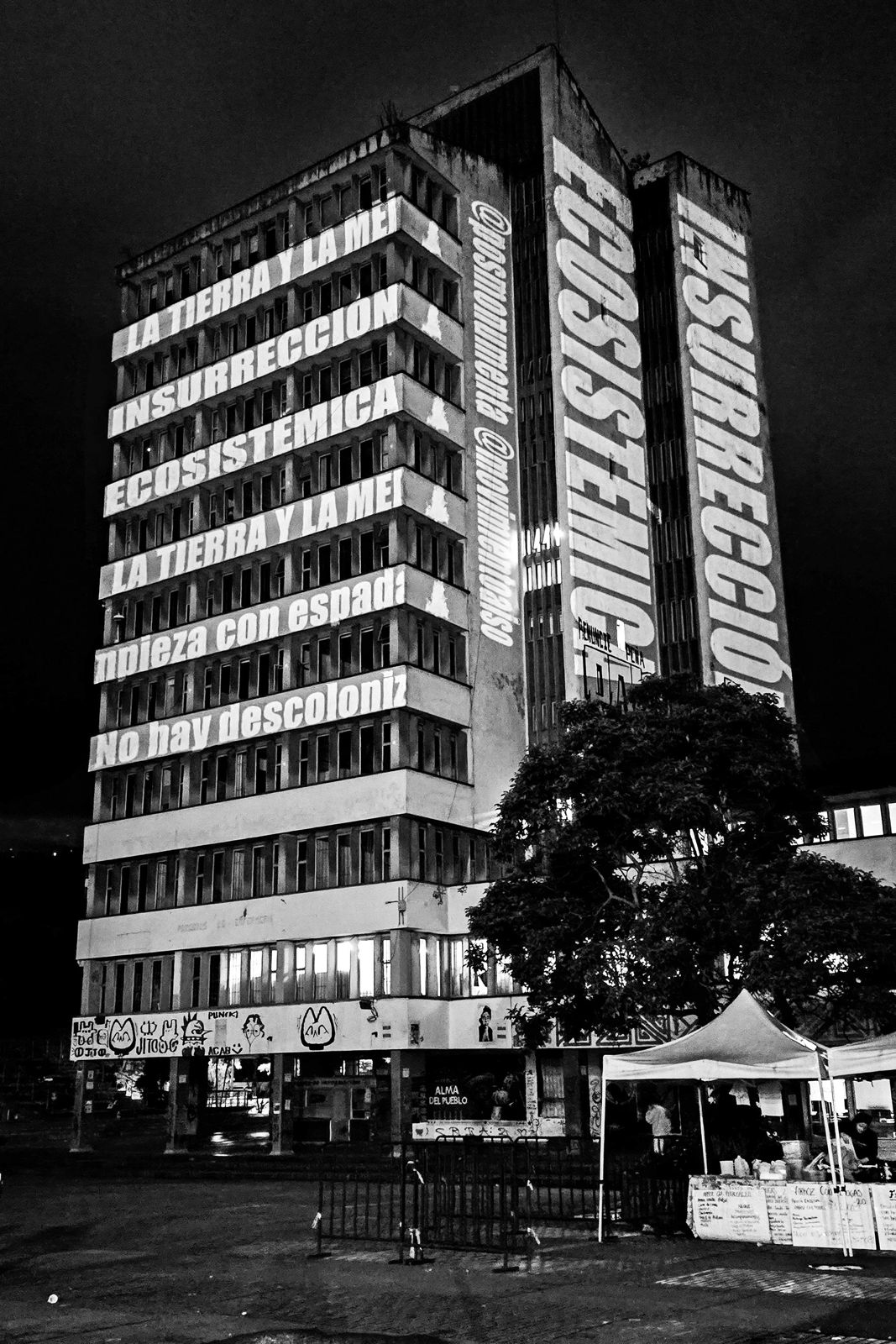 Insurrección Ecosistémica de Julián Santana Rodríguez. En el contexto de la exposición «Imágenes de la Resistencia» realizada en la Universidad Nacional, el colectivo Posmonumenta activó el espacio público de la Plaza Che el 10 de junio de 2025 con una acción de videomapping tipográfico. Sobre la fachada de la Torre de Enfermería se proyectaron mensajes de resistencia medioambiental, testimonios y manifiestos de re-existencia ecosistémica y comunitaria, mientras un Círculo de Palabra y Escucha se desarrollaba en simultáneo, tejiendo un diálogo vivo entre la imagen proyectada y las voces presentes.
