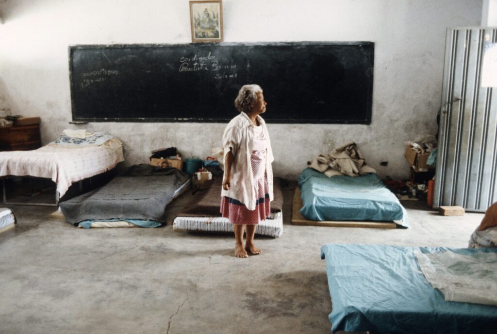 Mujer, adulta mayor, de pie y descalza frente a colchonetas en el piso. Quienes no vivían en tiendas de campaña, lo hacían en salones de clase.